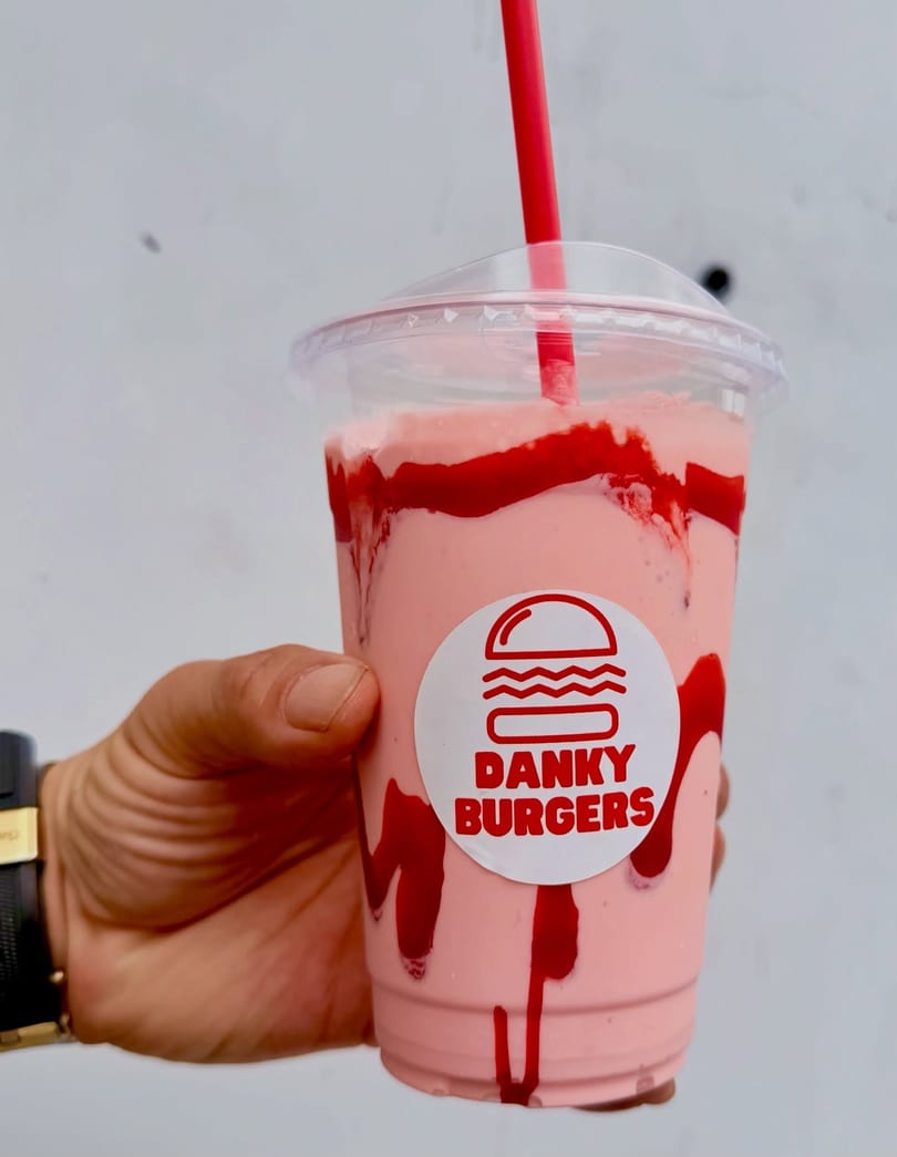 Hand holding a Danky Burgers strawberry milkshake with red syrup streaks and a red straw.