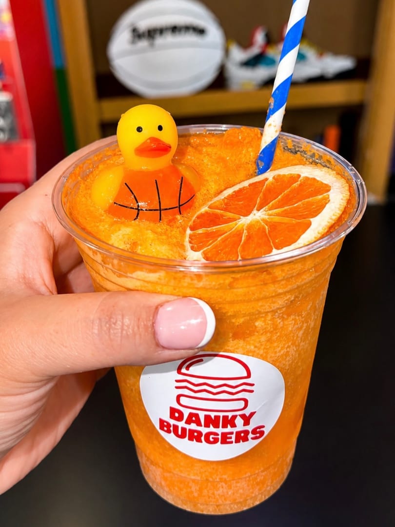 Hand holding a DANKY BURGERS orange drink with a rubber duck, orange slice, and blue straw.